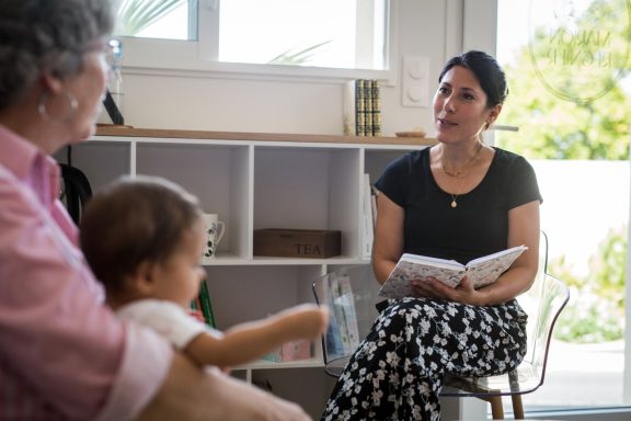 Une femme lit un livre à deux personnes, dont un enfant dans les bras d'un adulte.